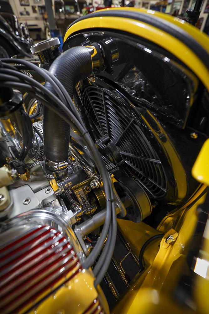 Close-up of the front radiator electric fan assembly, upper radiator hose, spark plug wires, and yellow engine bay frame rails.