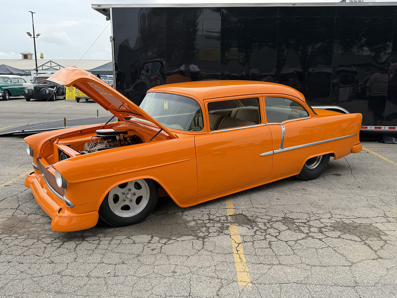 Side view of a smoothed, bright orange 1955 Chevrolet pro street car with hood propped open, aluminum wheels, and tucked rear slicks in front of an enclosed trailer.