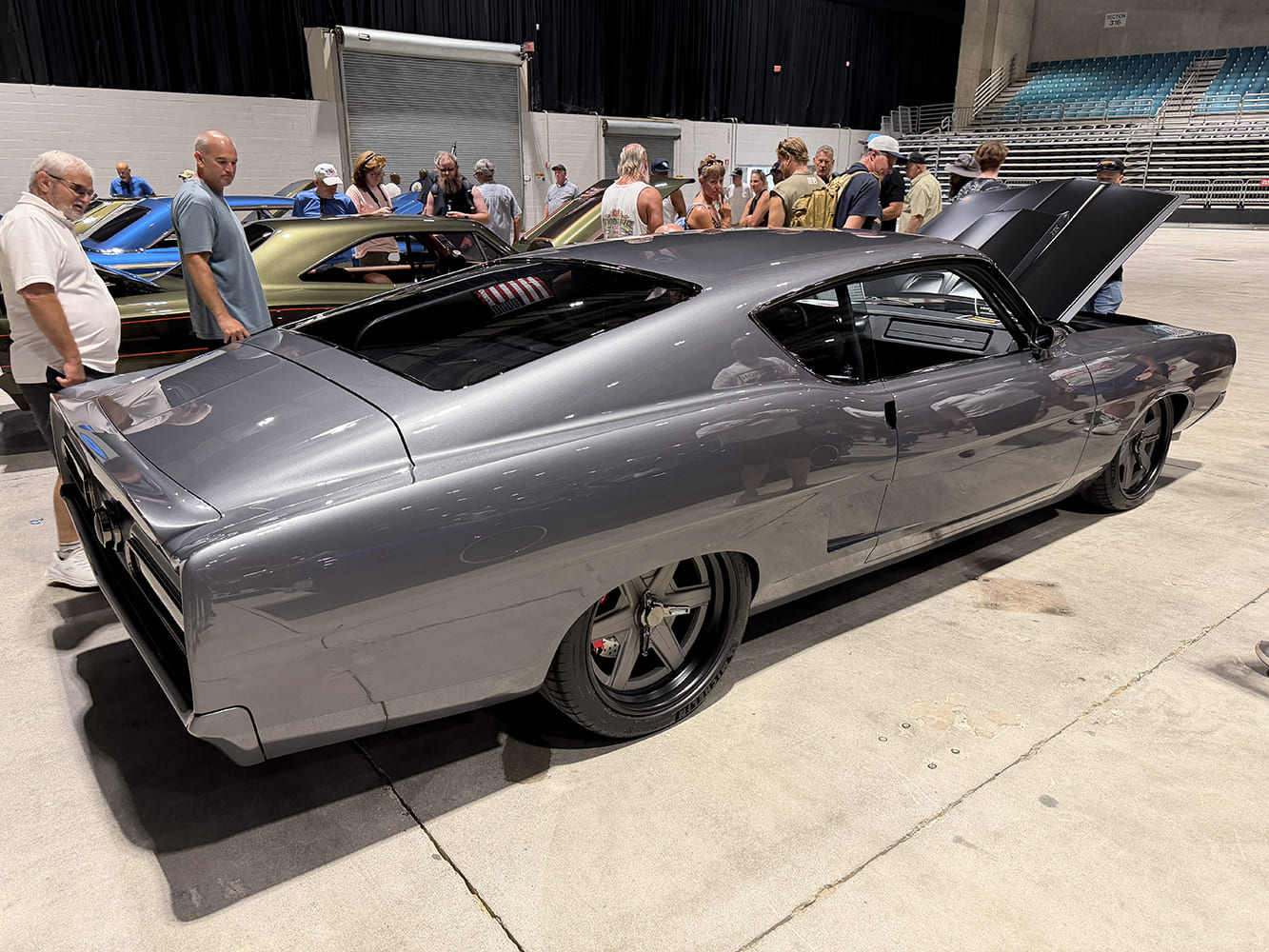 Rear three-quarter perspective of a pro-touring metallic charcoal 1969 Ford Torino showing custom wide black wheels, flush rear glass, and open hood in an exhibition hall.
