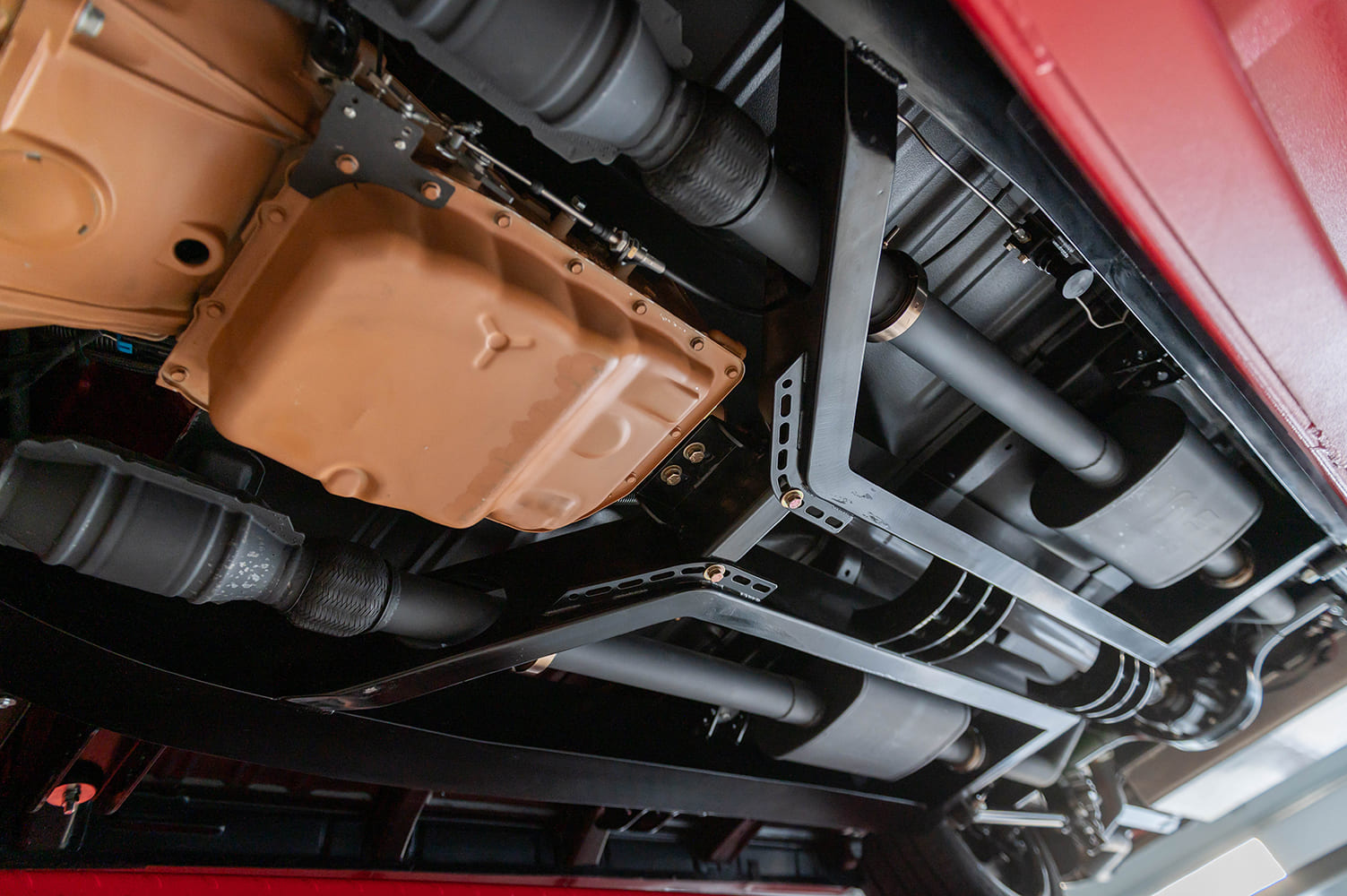 Looking forward along the underside of the floor pan showing the tan-painted transmission oil pan, tubular chassis crossmember, catalytic converters, and mufflers.