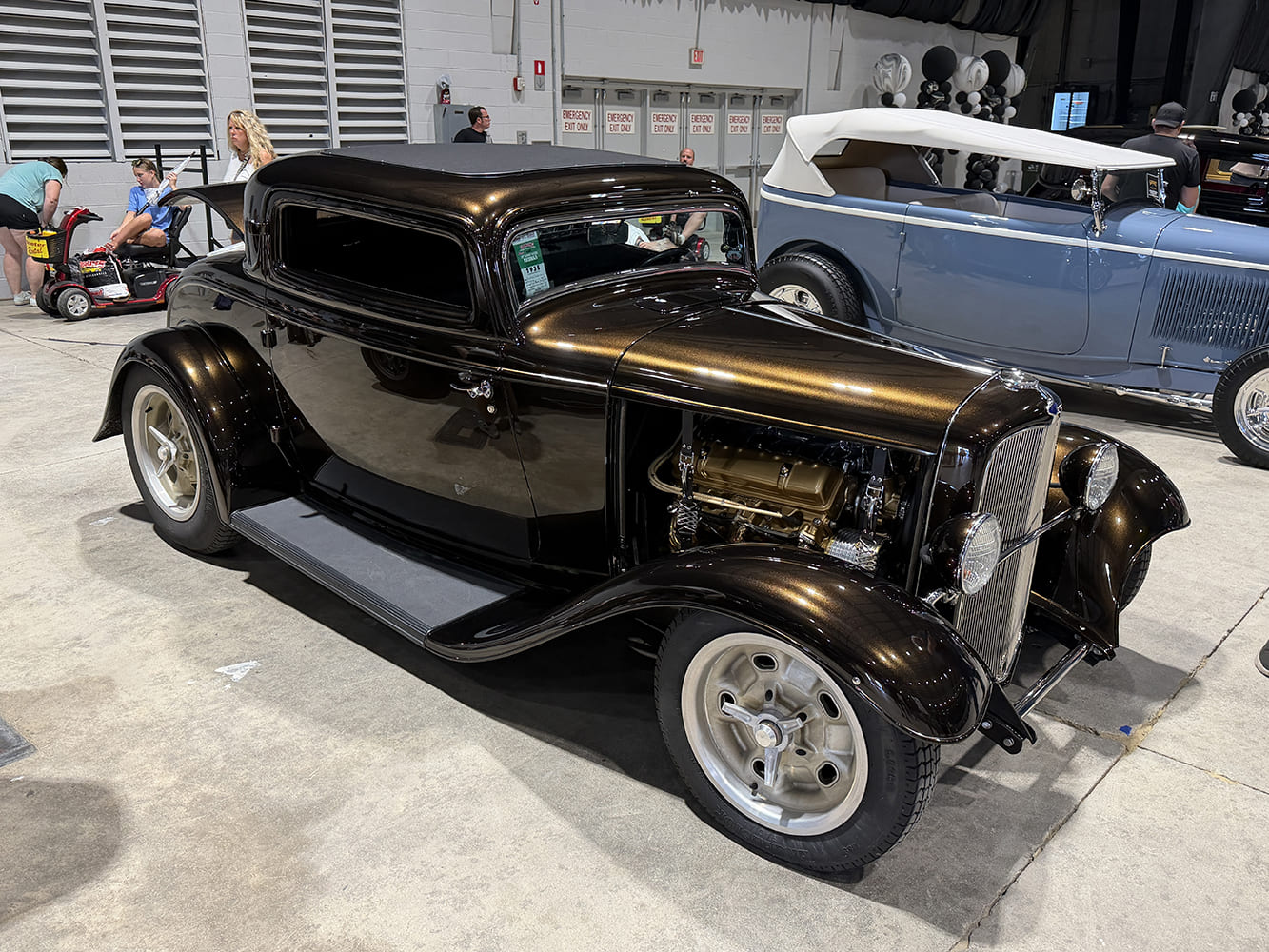 Front three-quarter shot of a deep root beer brown metallic 1932 Ford 3-window coupe hot rod with an exposed small-block V8 and Halibrand-style wheels indoors.