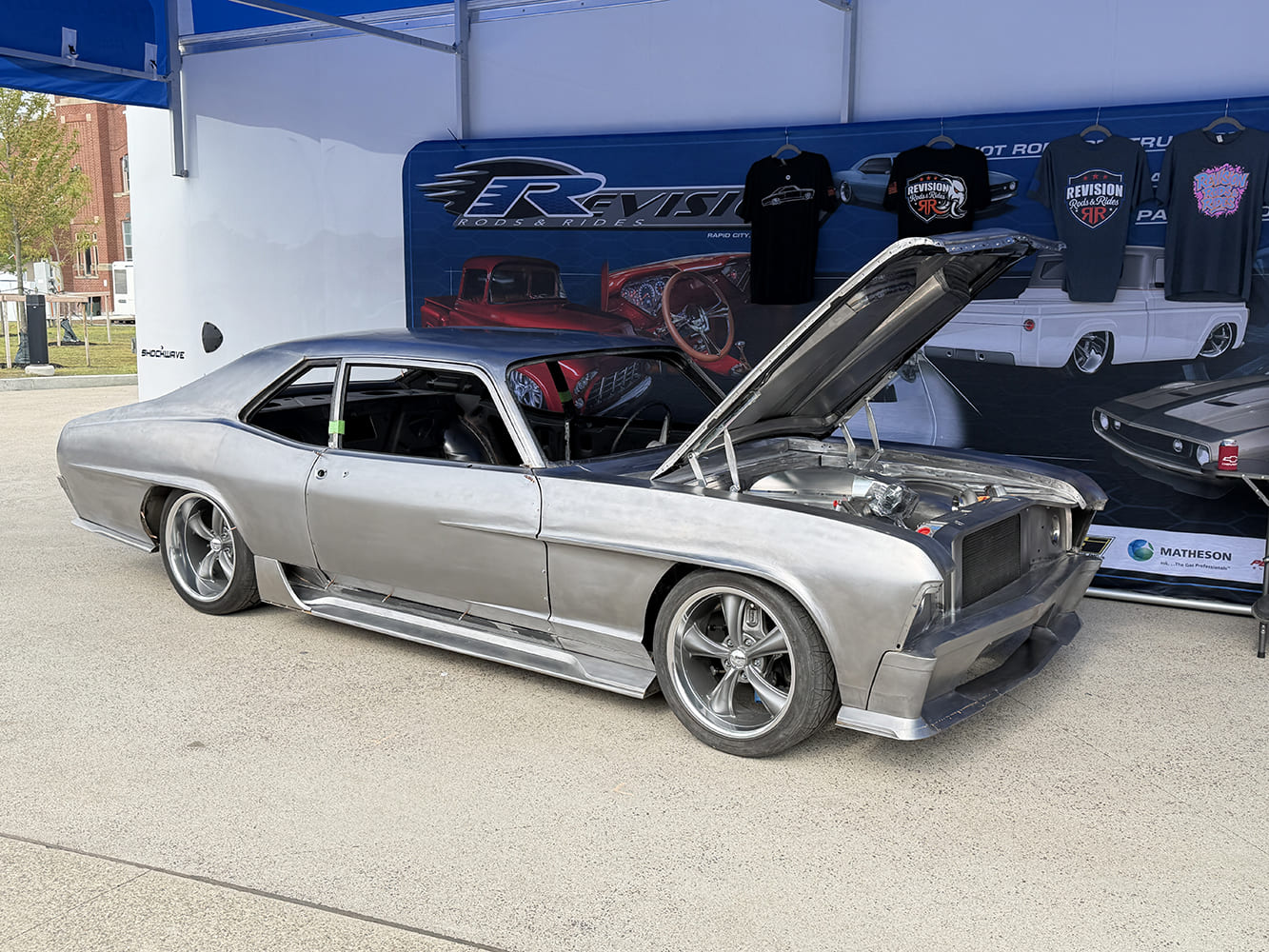 Side three-quarter view of a bare sheetmetal 1970 Chevrolet Nova pro-touring project car with open hood displayed at the Revision Rods & Rides outdoor booth.