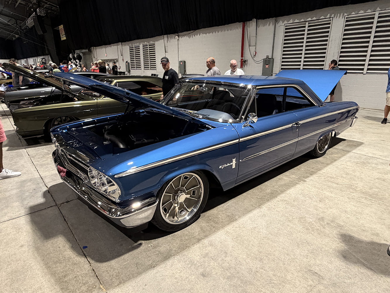 Front three-quarter angle of a metallic blue 1963 Ford Galaxie 500 with modern large-diameter polished turbine wheels and open hood inside the indoor showroom.