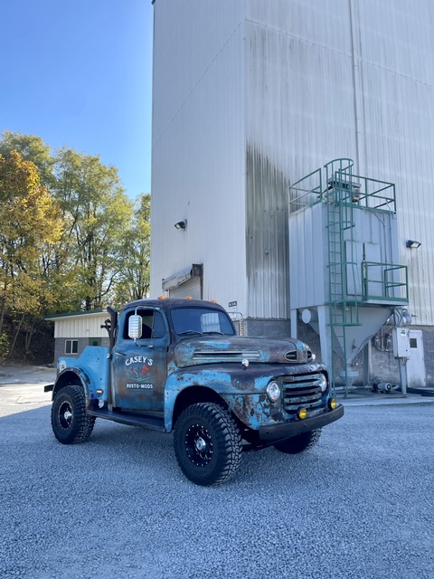 Driver-side perspective of the lifted patina blue 1949 Ford F2 wrecker conversion parked on gravel beside a tall white industrial building and silo.