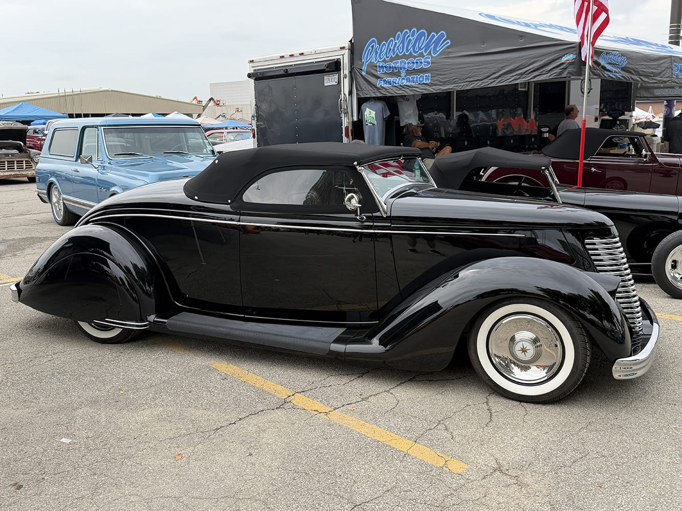 Side profile of a swept-back, black custom 1936 Ford convertible with smooth fender skirts, black canvas top, and chrome dish hubcaps parked by the vendor tent.