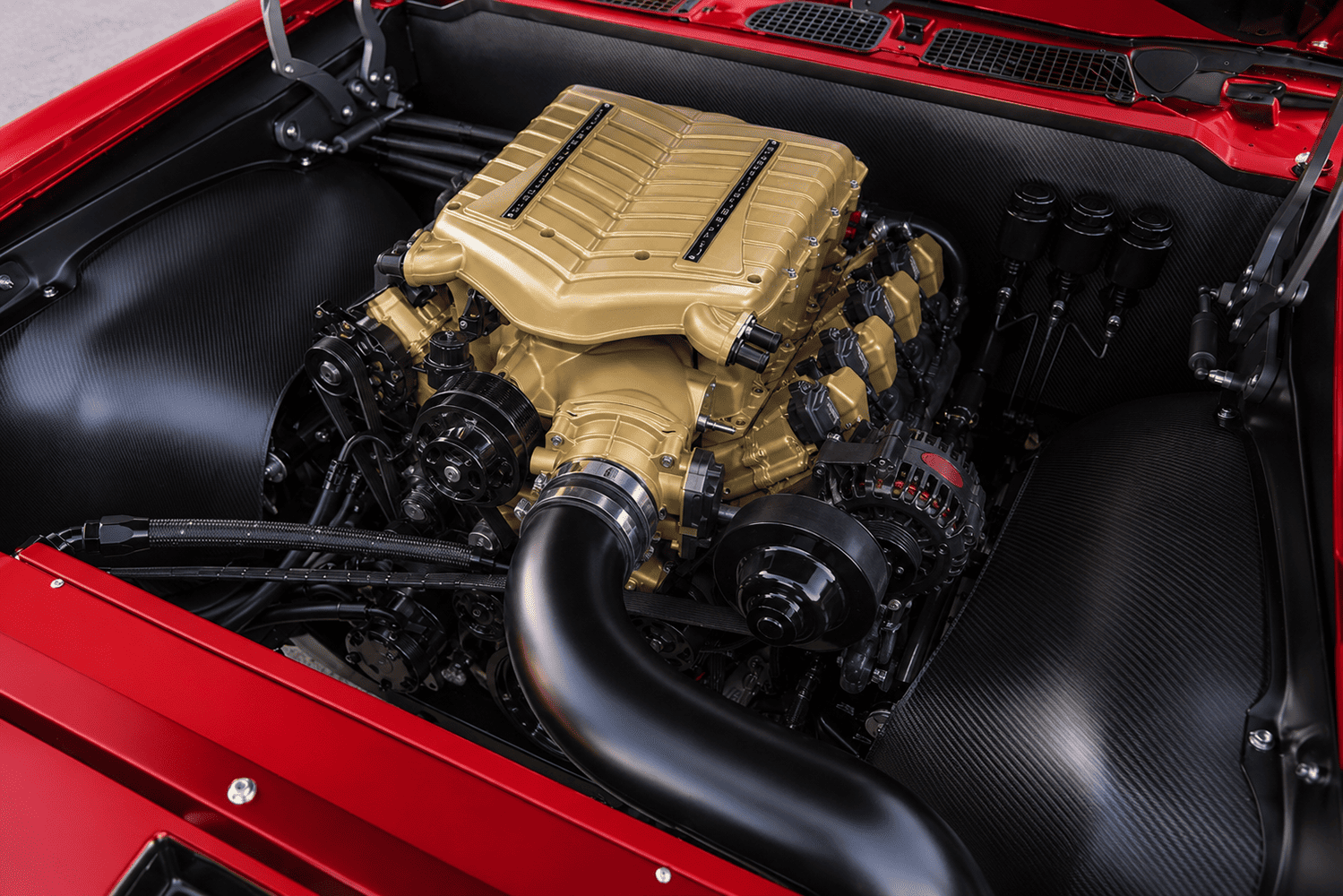 High-angle engine bay shot highlighting a gold-finished supercharged GM L8P crate engine framed by gloss carbon-fiber inner wheel tubs and black firewall.