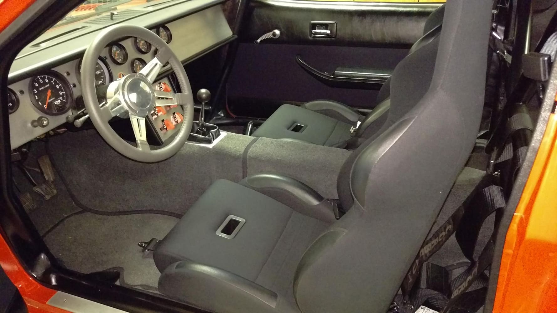 Interior view through the driver door showing the custom aluminum dashboard panel filled with round analog gauges, three-spoke steering wheel, and Doug Nash five-speed manual shifter.