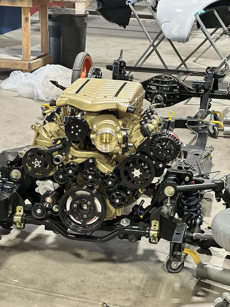 Front view of a gold powder-coated GM L8P engine with Whipple supercharger and black accessory drive mounted onto a bare Roadster Shop chassis on shop dollies.