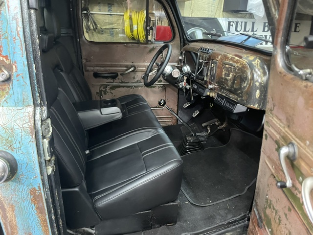 Open-door interior view of the 1949 Ford cab showing modern black leather seats with center console, manual floor shifter, and weathered patina dashboard.