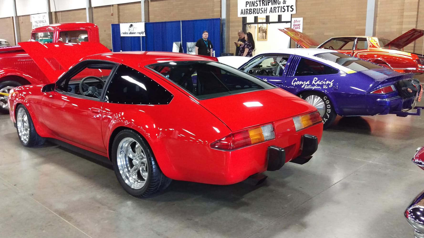 Rear three-quarter view of the bright red 1975 Chevy Monza on polished American mag wheels displayed on the indoor convention center floor.