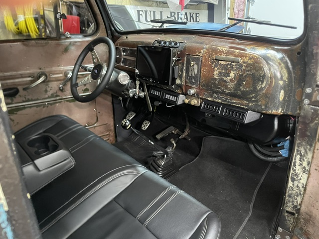 Close-up of the truck's cockpit featuring a large touchscreen infotainment display, under-dash air conditioning vents, and an aftermarket steering wheel mounted to a weathered steel dash.