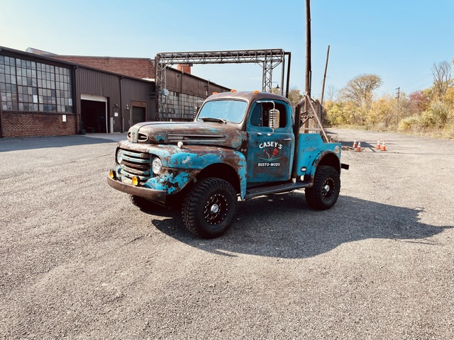 Full exterior perspective of the blue and rust patina 1949 Ford F2 4x4 parked in an open gravel lot outside a brick warehouse facility.