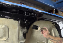 Hand using a paintbrush to spread thick gloss black rust encapsulator paint over the inner firewall and cowl cavity of a Chevrolet C10 cab mounted on a rolling stand.