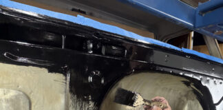 Hand using a paintbrush to spread thick gloss black rust encapsulator paint over the inner firewall and cowl cavity of a Chevrolet C10 cab mounted on a rolling stand.