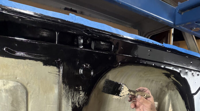 Hand using a paintbrush to spread thick gloss black rust encapsulator paint over the inner firewall and cowl cavity of a Chevrolet C10 cab mounted on a rolling stand.