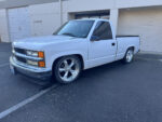 1988-1991 Chevy OBS Gauges – Dakota Digital’s RTX System Front three-quarter view of a lowered white 1988–1994 Chevrolet C1500 regular cab pickup on chrome five-spoke wheels parked outside commercial roll-up doors.