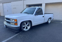 Front three-quarter view of a lowered white 1988–1994 Chevrolet C1500 regular cab pickup on chrome five-spoke wheels parked outside commercial roll-up doors.