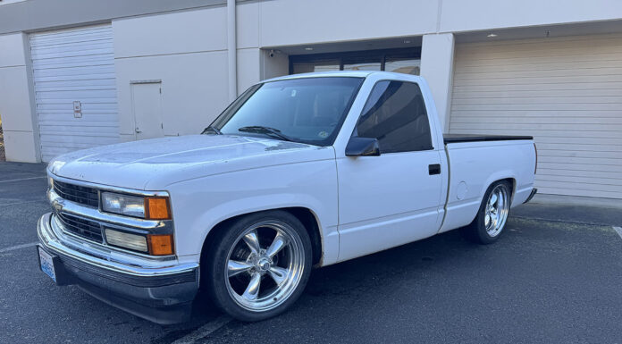 Front three-quarter view of a lowered white 1988–1994 Chevrolet C1500 regular cab pickup on chrome five-spoke wheels parked outside commercial roll-up doors.