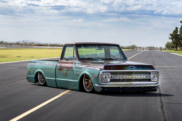 Low-angle front three-quarter view of a bagged patina turquoise and brown 1969 Chevrolet C10 pickup laid out on airport runway tarmac.