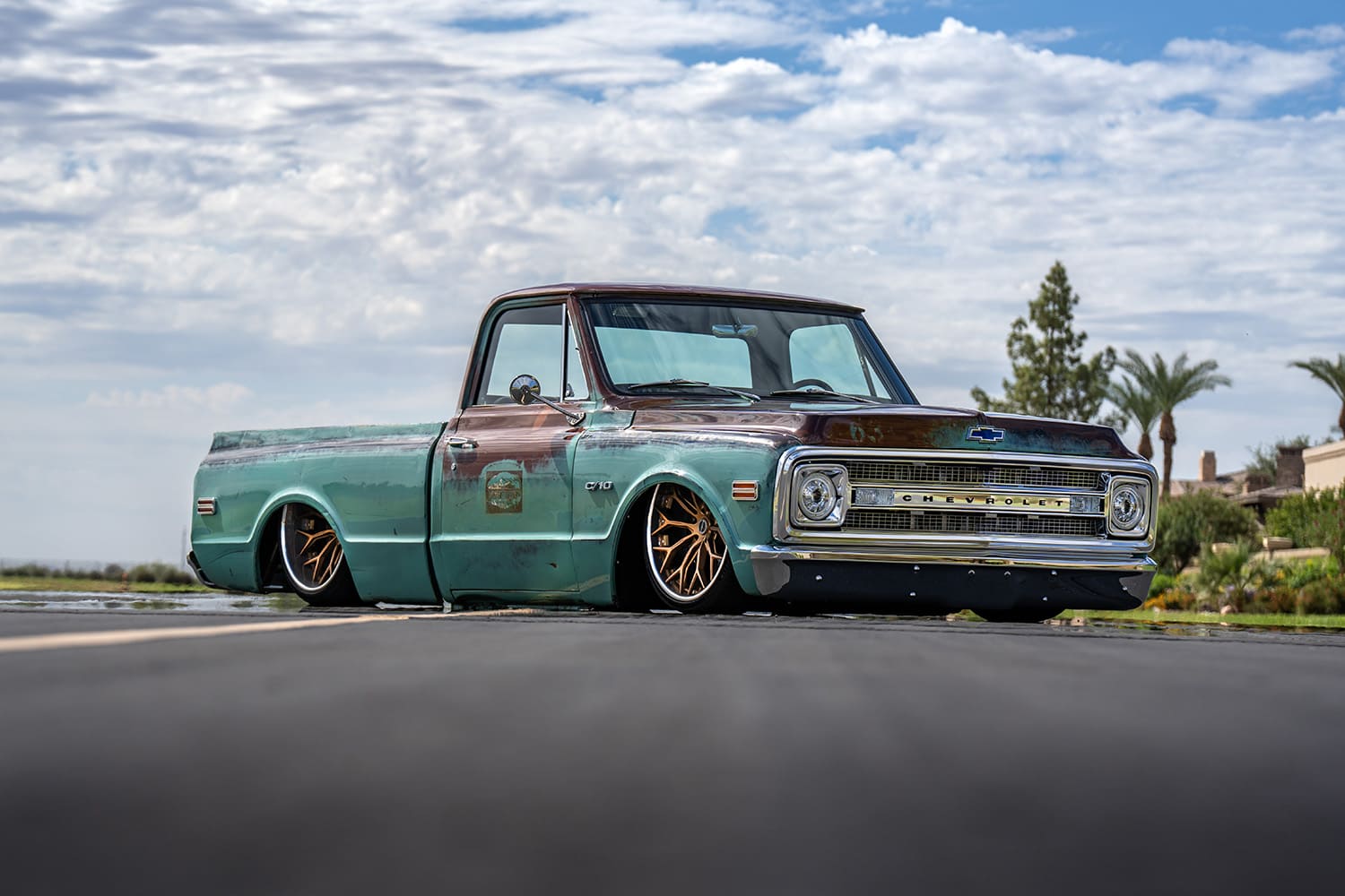 Extreme low-angle front three-quarter shot of the patina 1969 Chevy C10 showcasing its chrome grille, bumper, and custom bronze multi-spoke wheels.