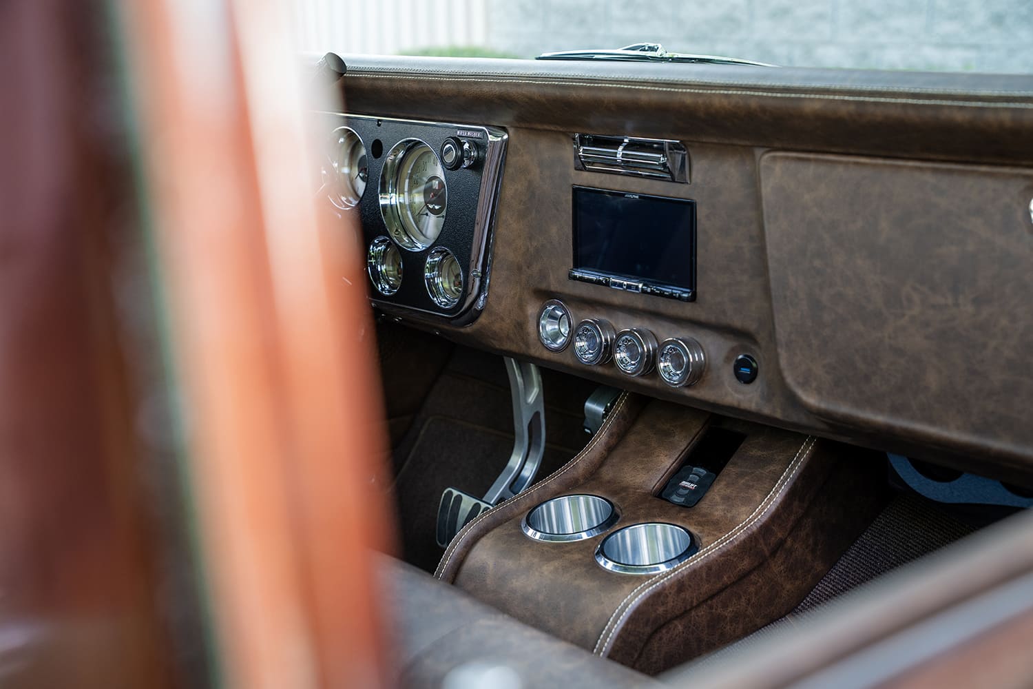 Passenger-side perspective of the distressed brown leather-wrapped dash pad, integrated touchscreen display, billet A/C vents, and cup holders.
