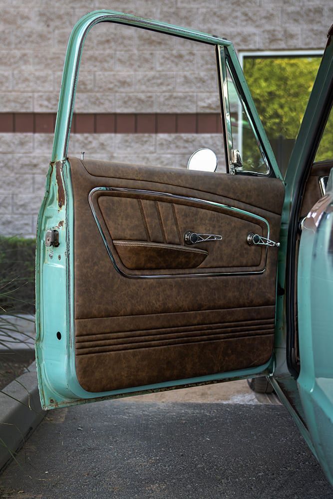 Open driver-side door showcasing a custom stitched brown leather door panel with chrome trim, matching armrest, and skeletonized billet handles against the turquoise jamb.