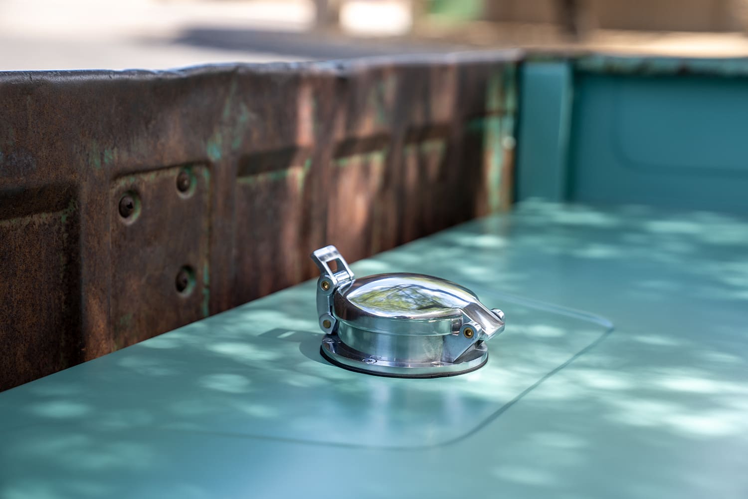 Macro view of a polished aluminum quick-fill fuel cap mounted flush into the turquoise sheetmetal bed floor in front of the weathered cab wall.