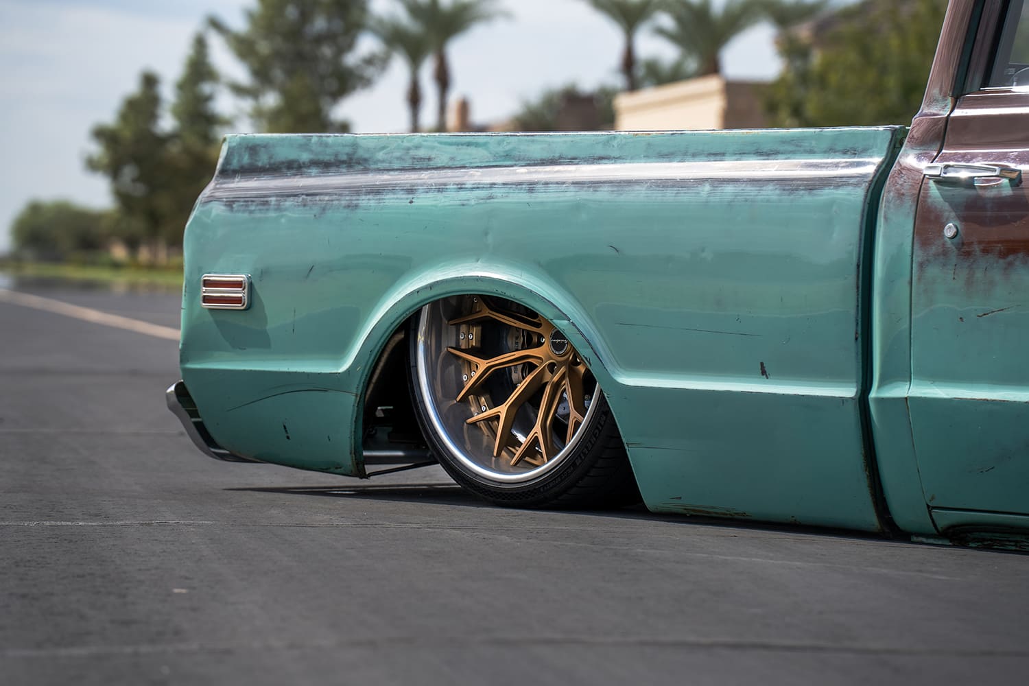 Low-angle rear quarter perspective showing the massive polished-lip bronze wheel deeply tucked into the faded turquoise bedside.