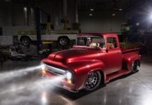 Front three-quarter studio view of a slammed candy brandywine 1956 Ford F100 pickup with projector headlights glowing brightly in a dark shop bay.