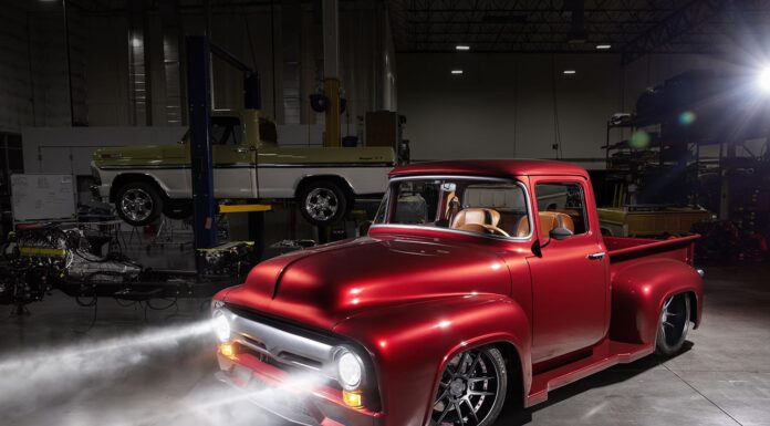 Front three-quarter studio view of a slammed candy brandywine 1956 Ford F100 pickup with projector headlights glowing brightly in a dark shop bay.