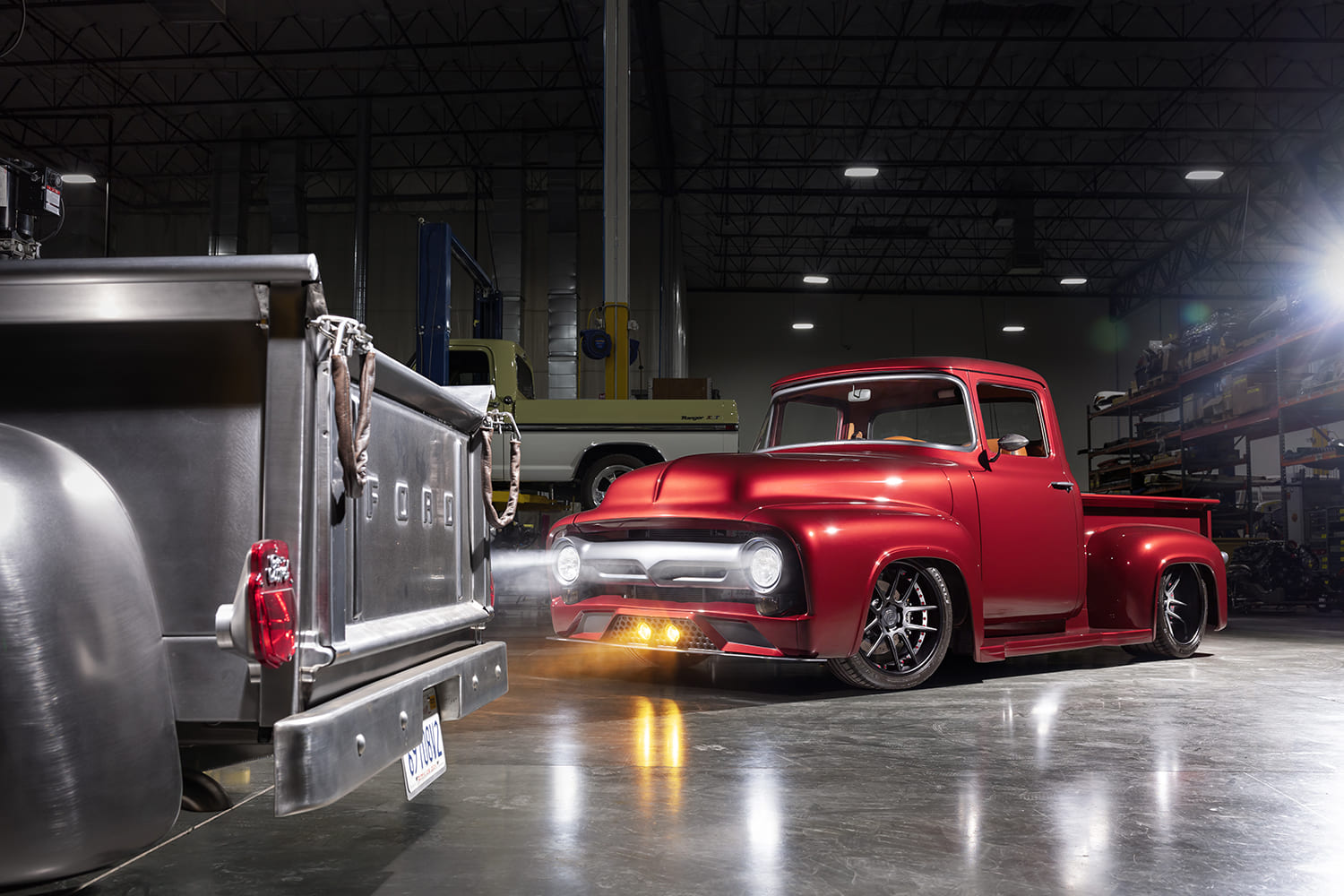 Wide workshop perspective showing the candy red 1956 Ford F100 beaming headlights across a polished concrete floor toward another classic truck bed.