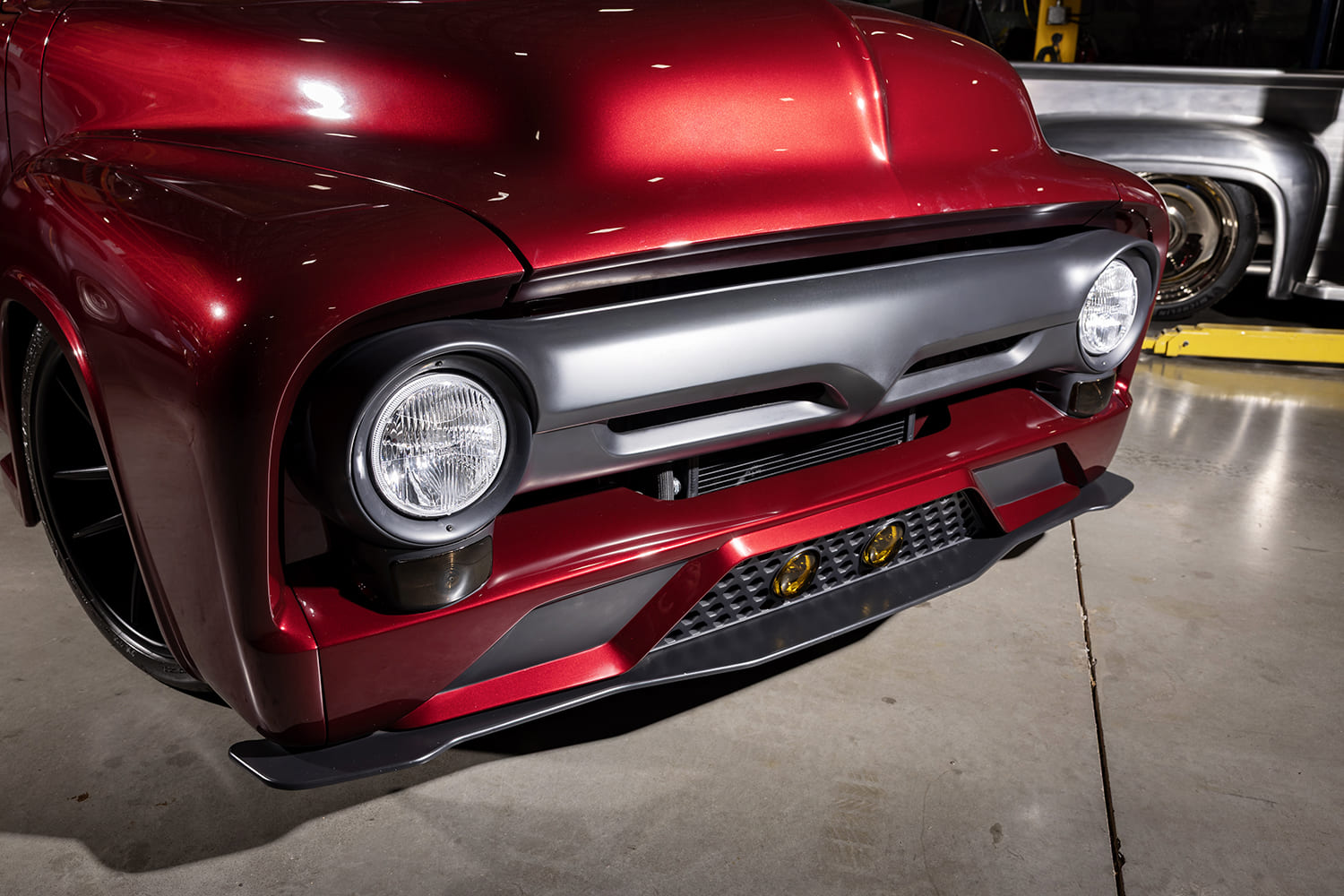 Close-up front three-quarter view showing the matte charcoal custom grille bar, modern round headlights, honeycomb lower air dam with amber lights, and chin splitter on the candy red 1956 Ford F100.