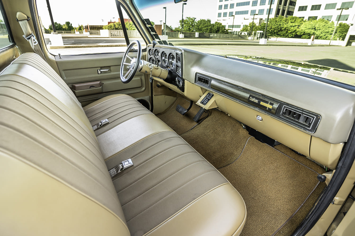Wide passenger-side view into the tan upholstered cab showing the factory-style split bench seat, billet steering wheel, and Dakota Digital instrument cluster.