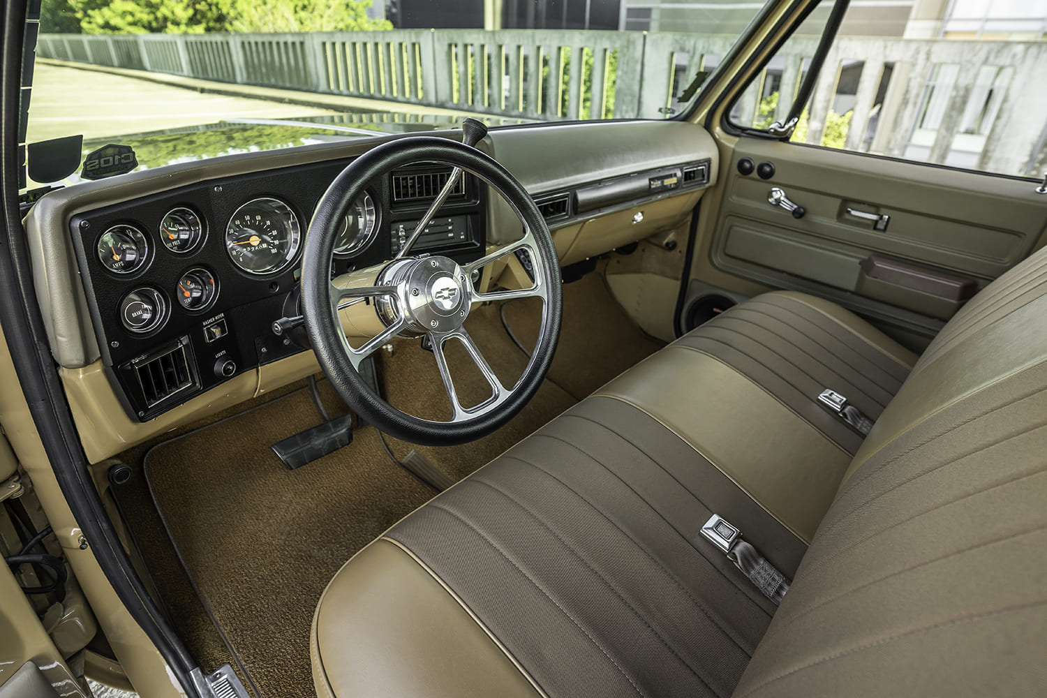 High three-quarter angle behind the polished four-spoke billet steering wheel overlooking the tan dashboard, Vintage Air vents, and loop carpet.