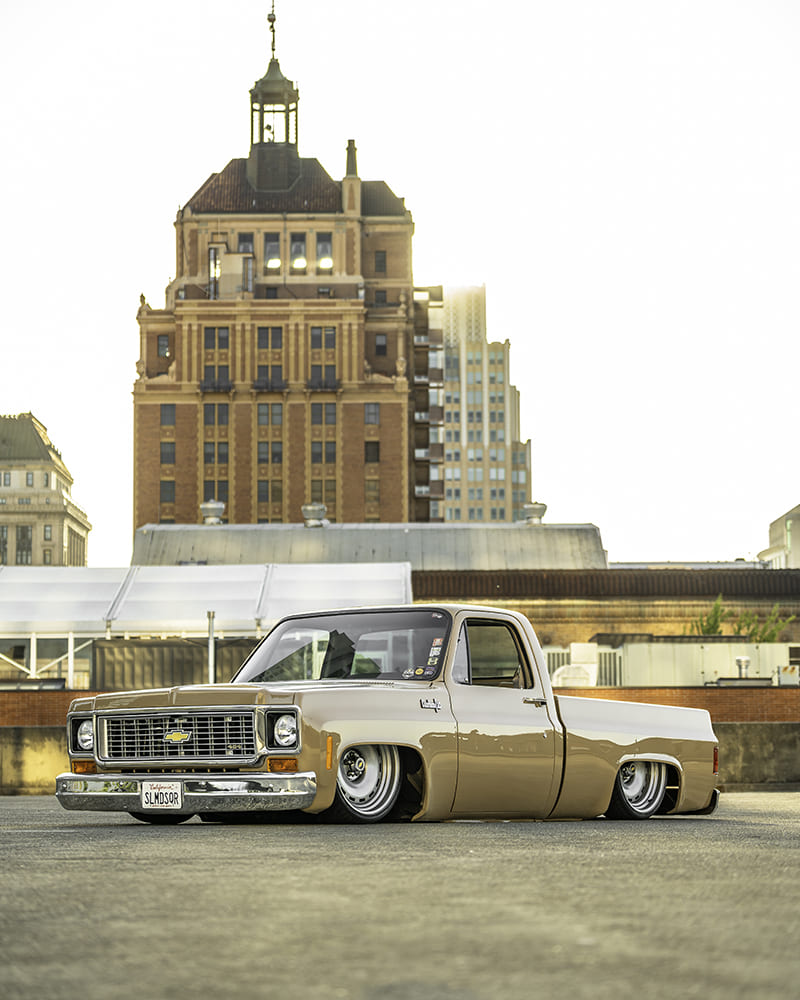 Vertical rooftop composition of the slammed 1973 Chevy Squarebody parked on asphalt in front of a historic multi-story brick building.