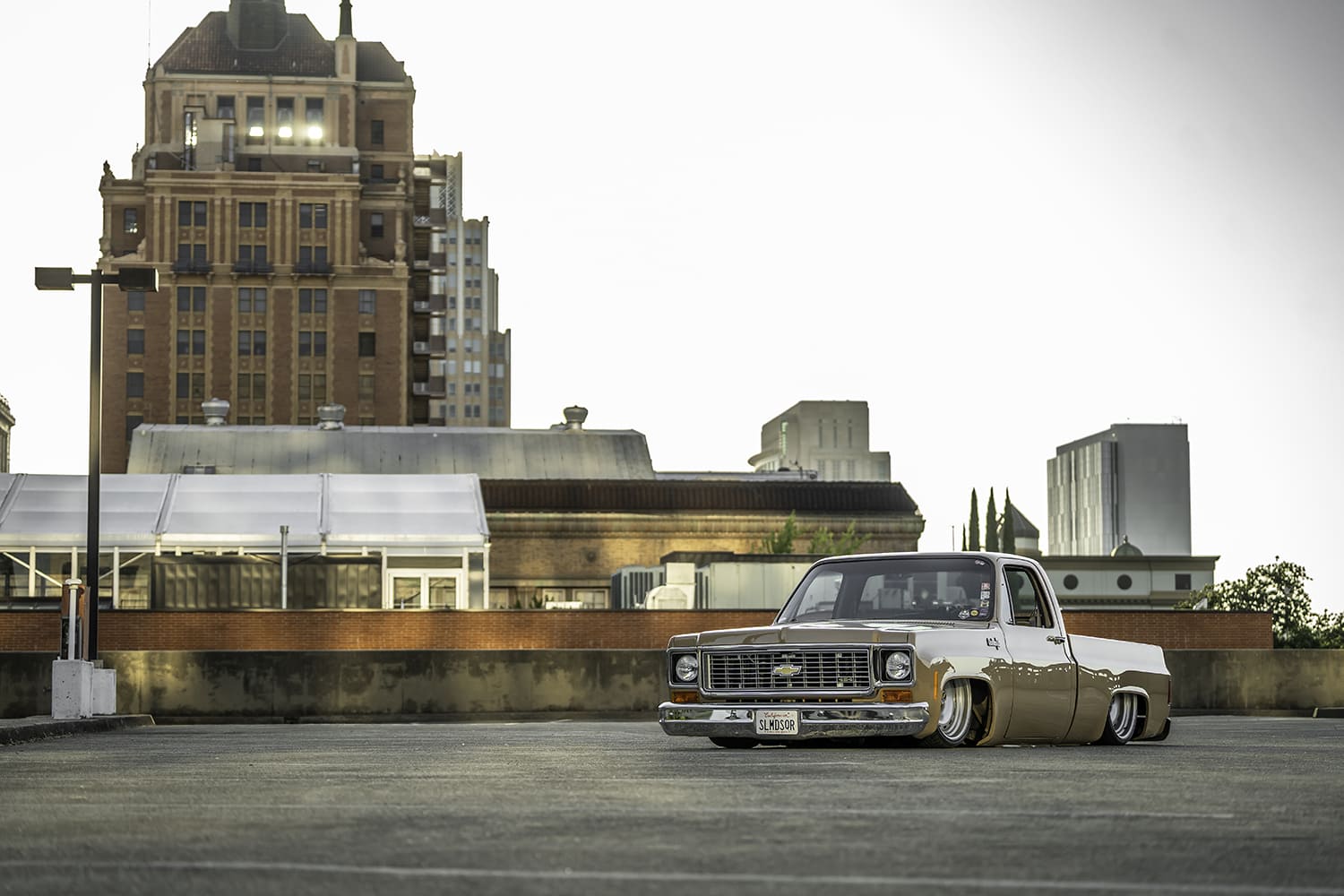 Wide-angle front three-quarter view of the slammed Mojave Tan 1973 Chevy C10 shortbed resting frame-flat on an open rooftop parking lot framed by urban architecture.